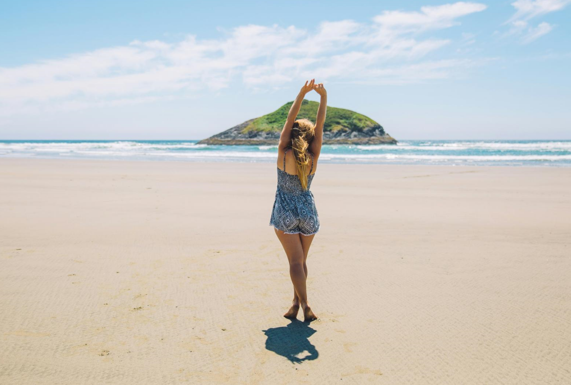 A woman standing on a sandy beach, stretching her arms above her head, suggesting freshness and confidence likely from using deodorant.